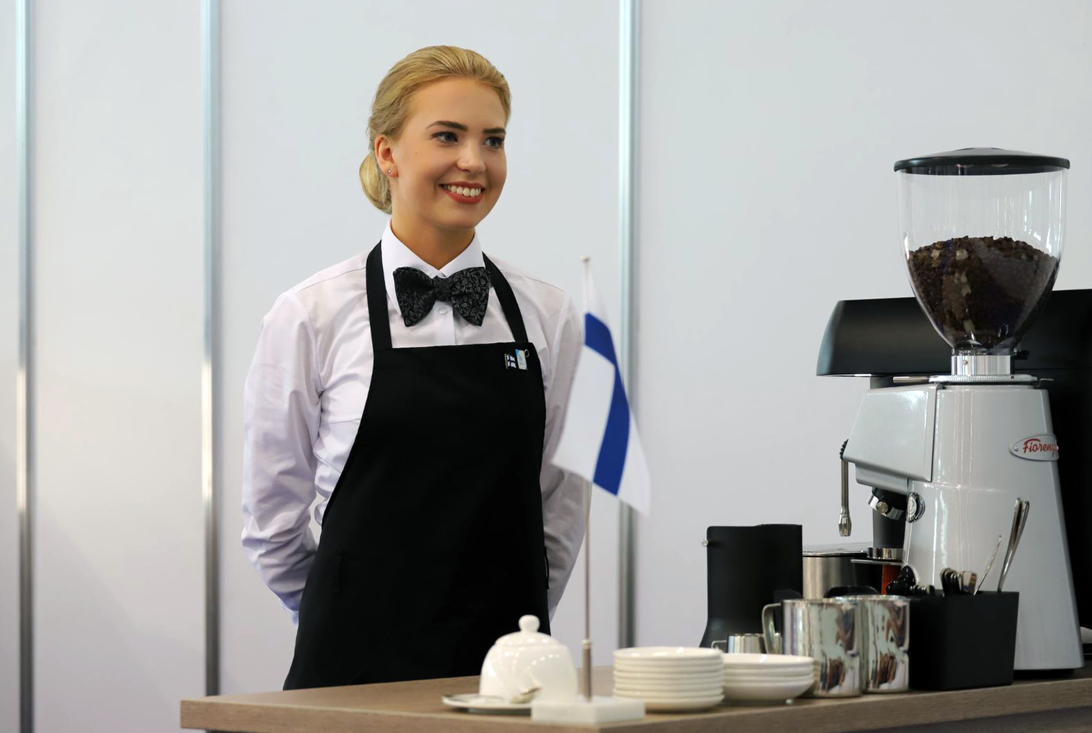 Nainen seisoo ja hymyilee kahvinkeittimen ja astioiden vieressä.