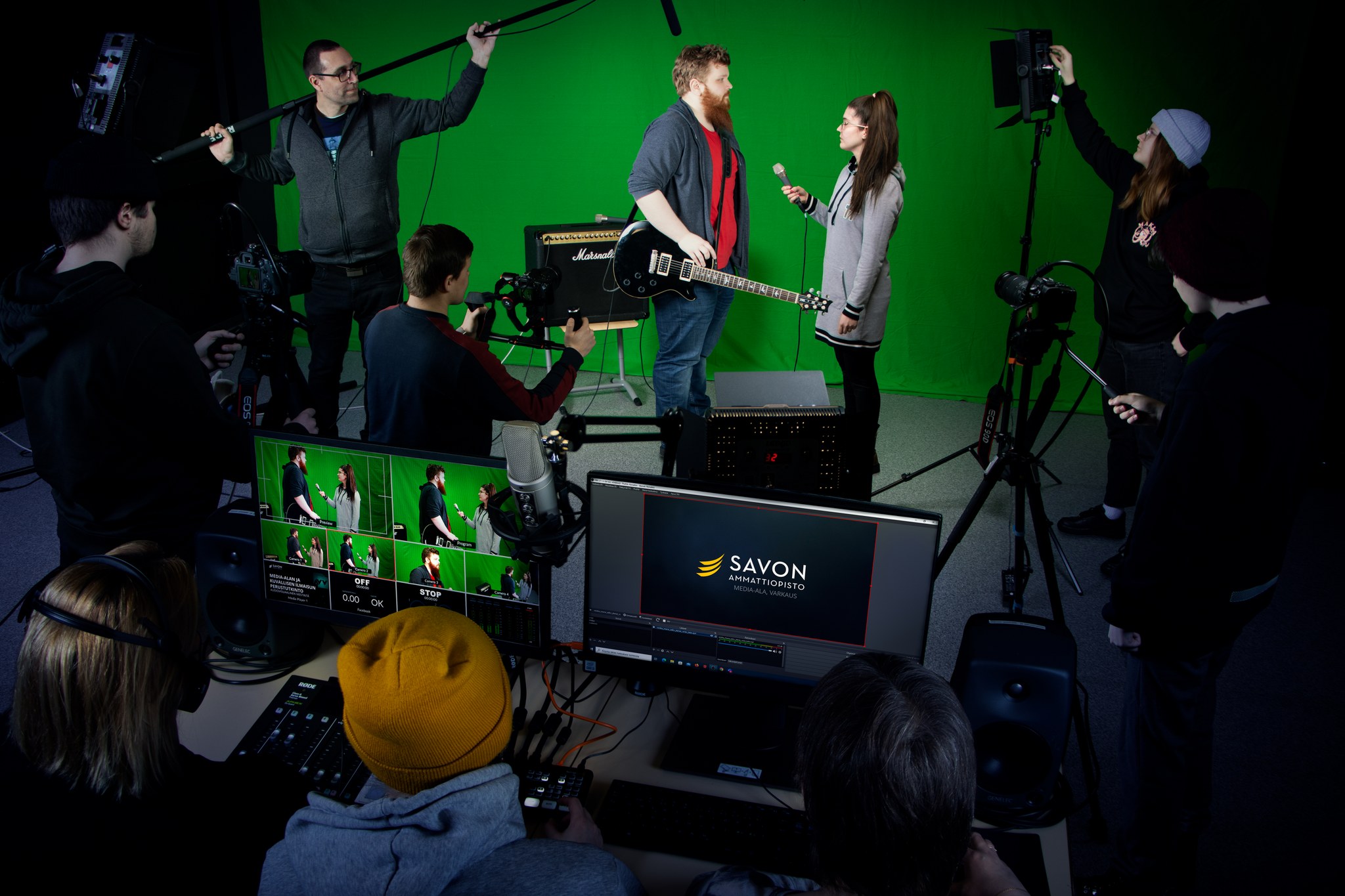 Behind the scene -kuva, jossa vihreää kangasta vasten on haastattelutilanne ja taustalla on valo- ja äänimiehet ja edessä kamerat.