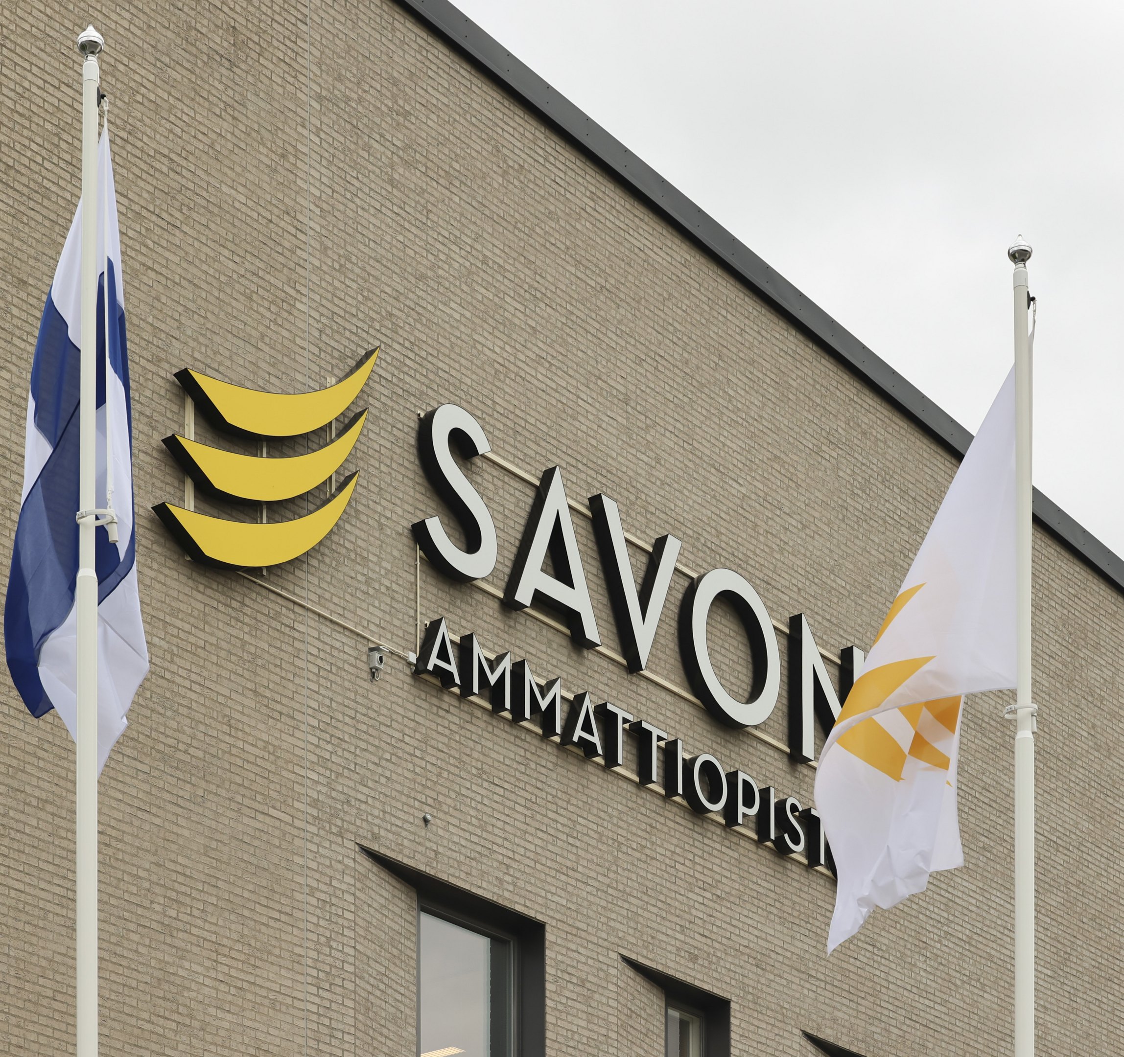 Savon ammattiopiston logo koulun seinässä ja ympärillä Suomen lippu ja Sakkyn lippu.