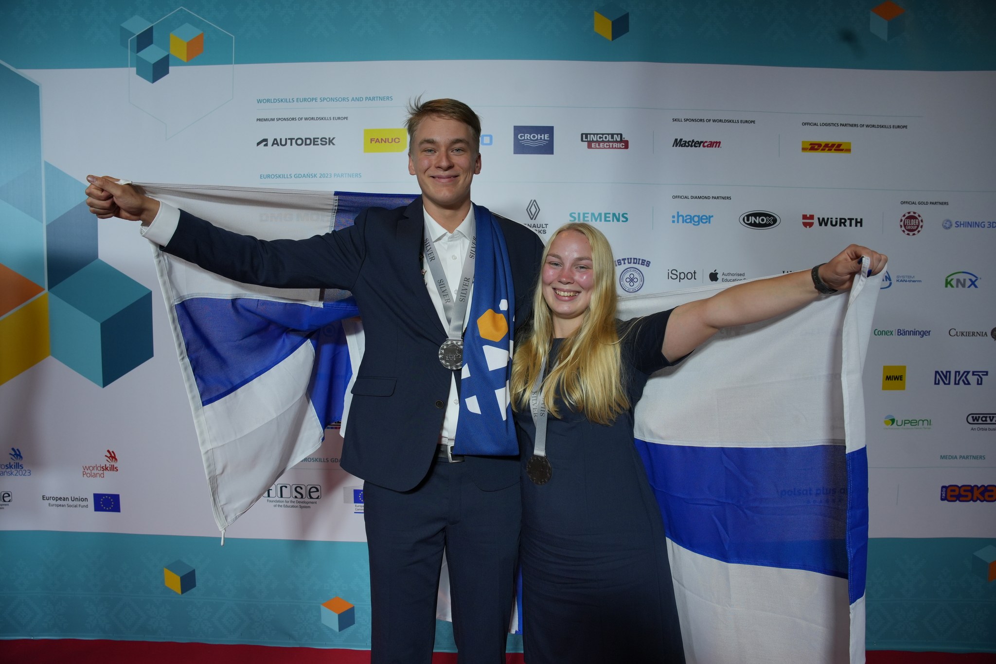 Kaksi nuorta hienoissa puvuissa pitävät Suomen lippua ja ovat iloisia.