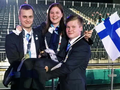 Nainen ja kaksi miestä hymyilevät. Nainen pitelee Suomen lippua.