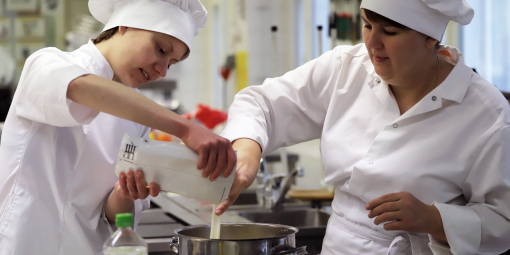 Kaksi naista valmistaa ruokaa elintarvikealan suojavaatteissa.