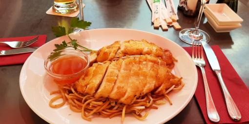 Ravintolan pöydällä lautasella on ruoka-annos, jossa on schnitzelin alla nuudeleita ja vasemmalla pienessä lasikupissa punaista kastiketta.