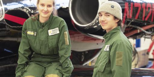 Kaksi vihreä haalarista opiskelijaa on lentokonehallissa lentokoneen edessä.