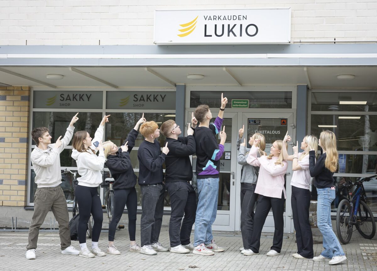Joukko opiskelijoita ulkona pääovien edessä osoittamassa Varkauden lukio kylttiä.