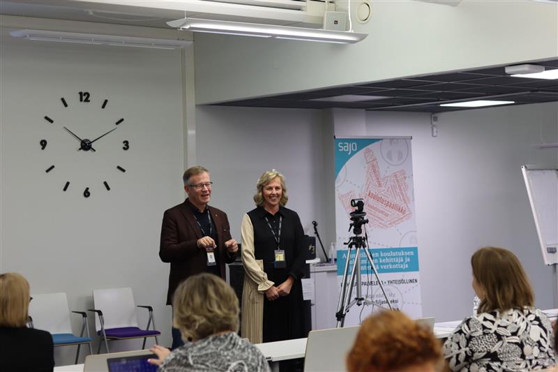 Kaksi ihmistä luokkatilan edessä ja joukko ihmisiä istuu. Kuva otettu takaa.