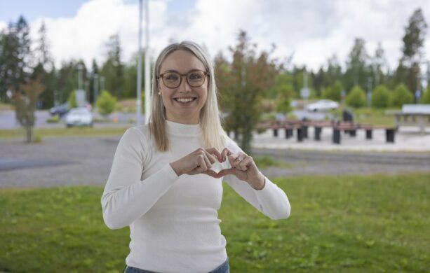 Ihminen hymyilee kameralle ja muodostaan sormillaan sydämen kuvan.