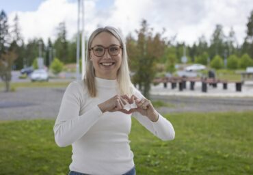 Ihminen näyttää käsillään sydämen merkin ja hymyilee kameralle.