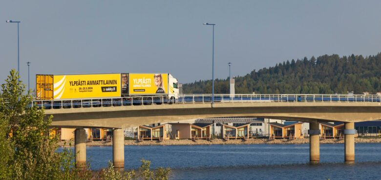 Yhdistelmäajoneuvo ajaa siltaa pitkin. Taivas ja vesi ovat sinisiä.
