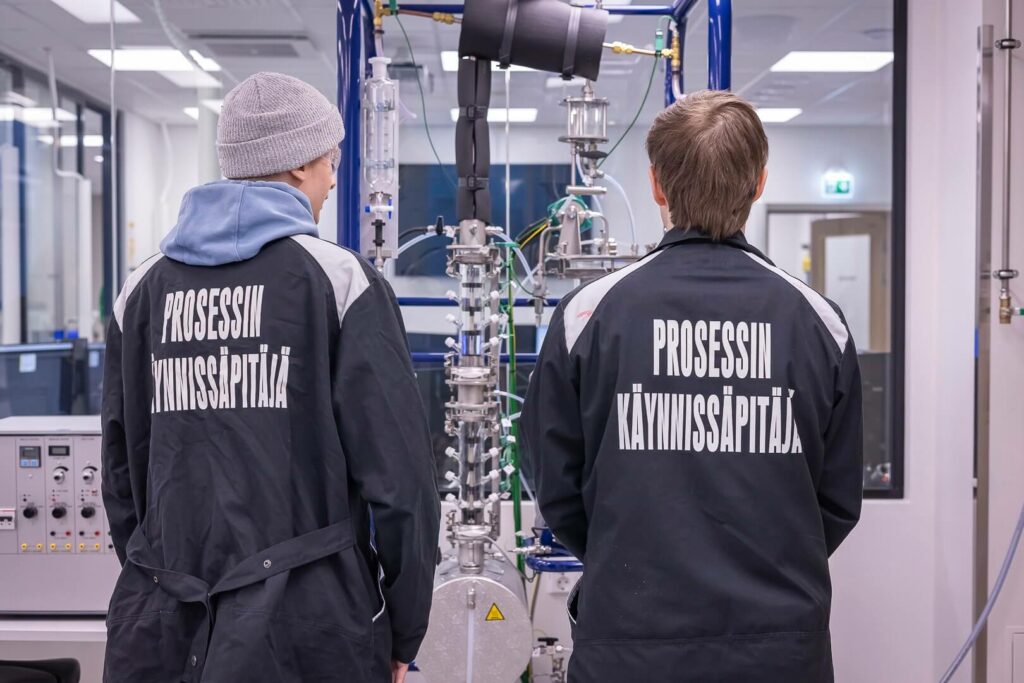 Kaksi henkilöä seisoo mittalaitteiston edessä ja selässä lukee prosessinhoitaja.