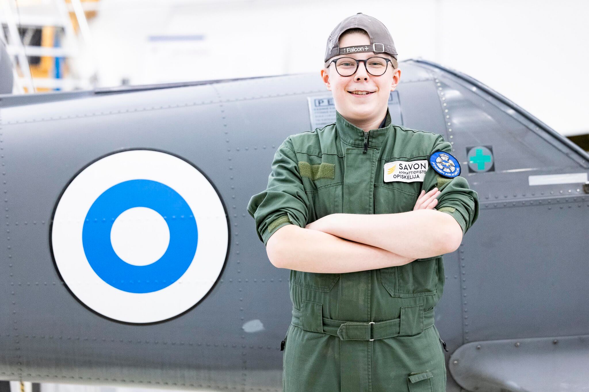 Lentokoneasentajaopiskelija lippalakki päässään hymyilee koneen edessä.