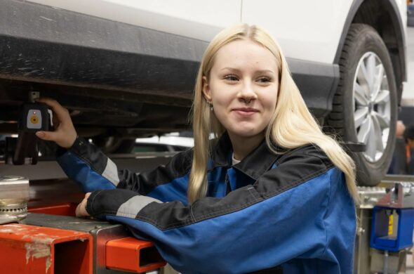 vaaleahiuksinen nainen seisoo auton vieressä työhaalarit yllään ja hymyilee