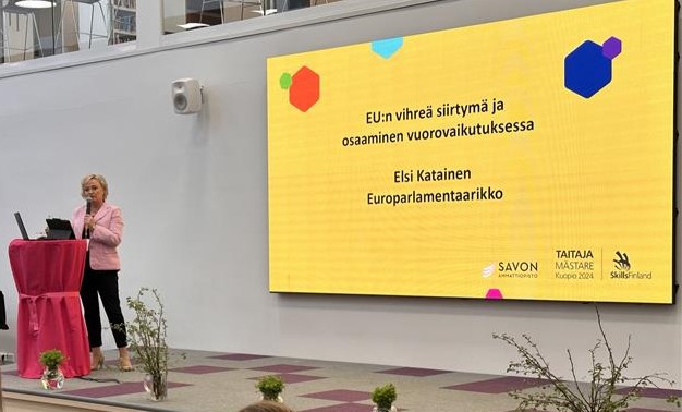Nainen seisoo lavalla ja puhuu mikrofoniin.