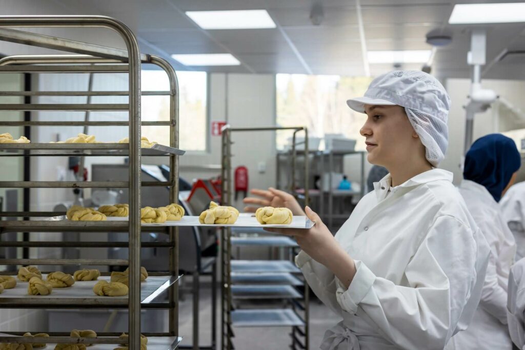 opiskelija laittaa korvapuusteja pellille ja uuniin. Yllä valkoinen työasu.