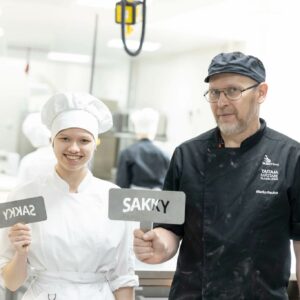 opettaja ja opiskelija seisovat leipomossa kyltit kädessään. Kyltissä lukee sakky.