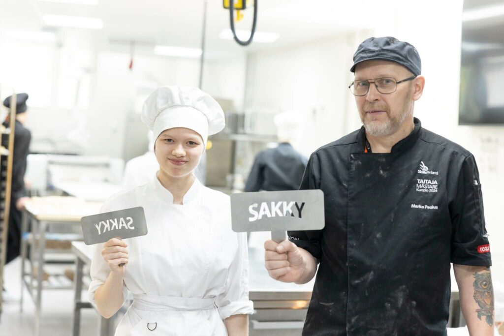 opettaja ja opiskelija seisovat leipomossa kyltit kädessään. Kyltissä lukee sakky.