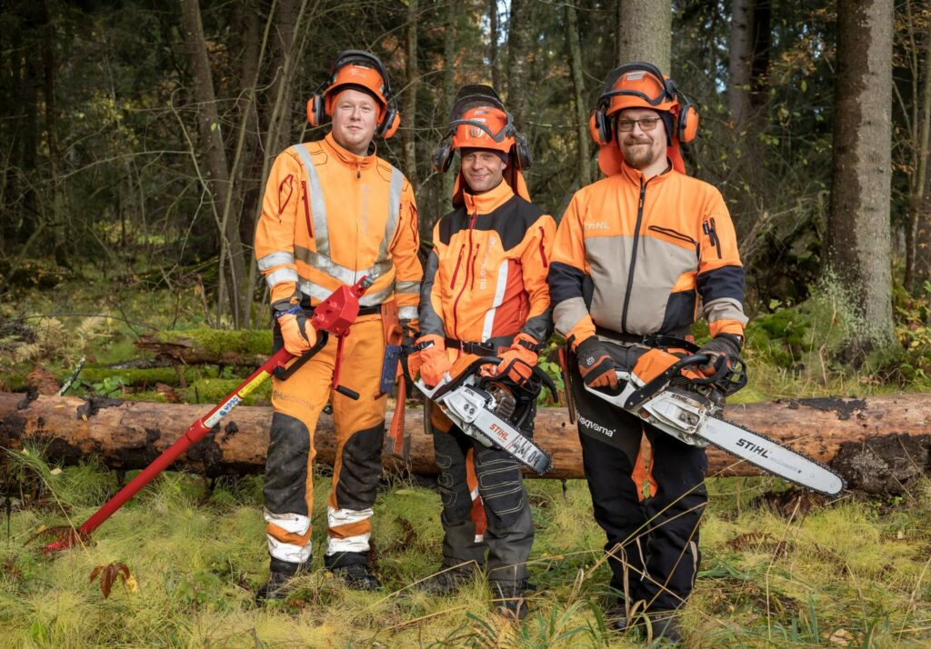 Kolme metsuria puun juurella ja kaikilla on moottorisahat tai muita työvälineitä käsissään.