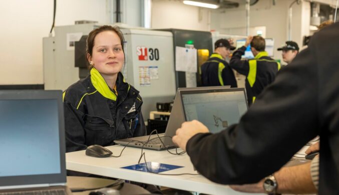 Nainen istuu ja pöydällä on tietokone.