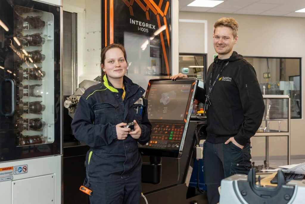 Kaksi henkilöä seisoo metallialan koneistuskoneen vieressä.