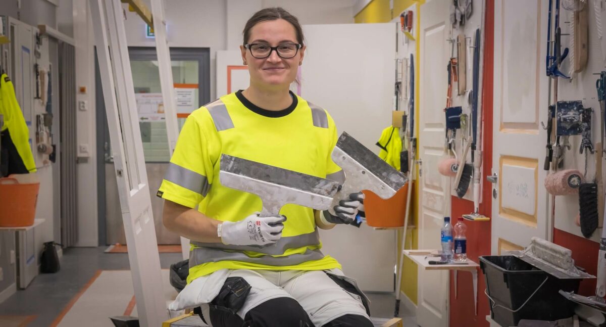 Henkilö istuu ja katsoo kameraan käsissään työvälineitä.