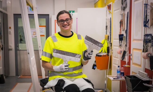 Henkilö katsoo kameraan ja hymyilee käsissään työvälneitä.