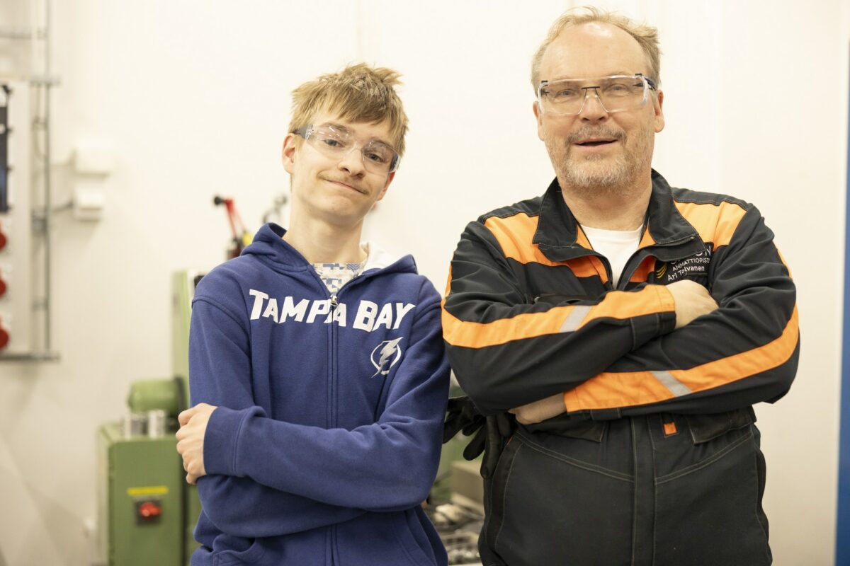 Opiskelija ja työhaalareissa oleva opettaja seisovat kädet puuskassa vierekkäin ja katsovat kameraan.