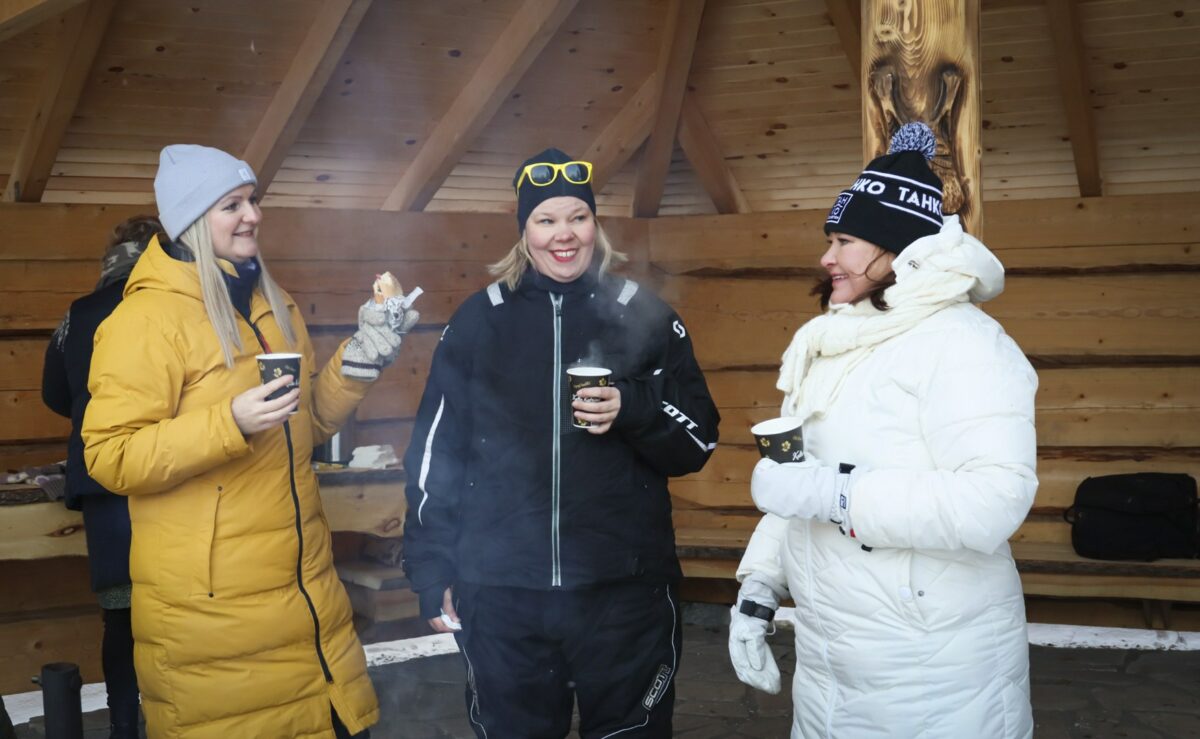 Kolme henkilöä laavulla, ja ihmisillä on pahviset kahvikupit kädessä.