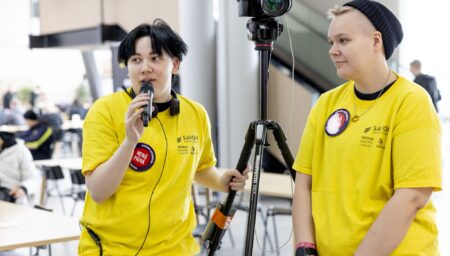 Keltapaitaisia opiskelijoita ja toinen puhuu mikrofoniin.