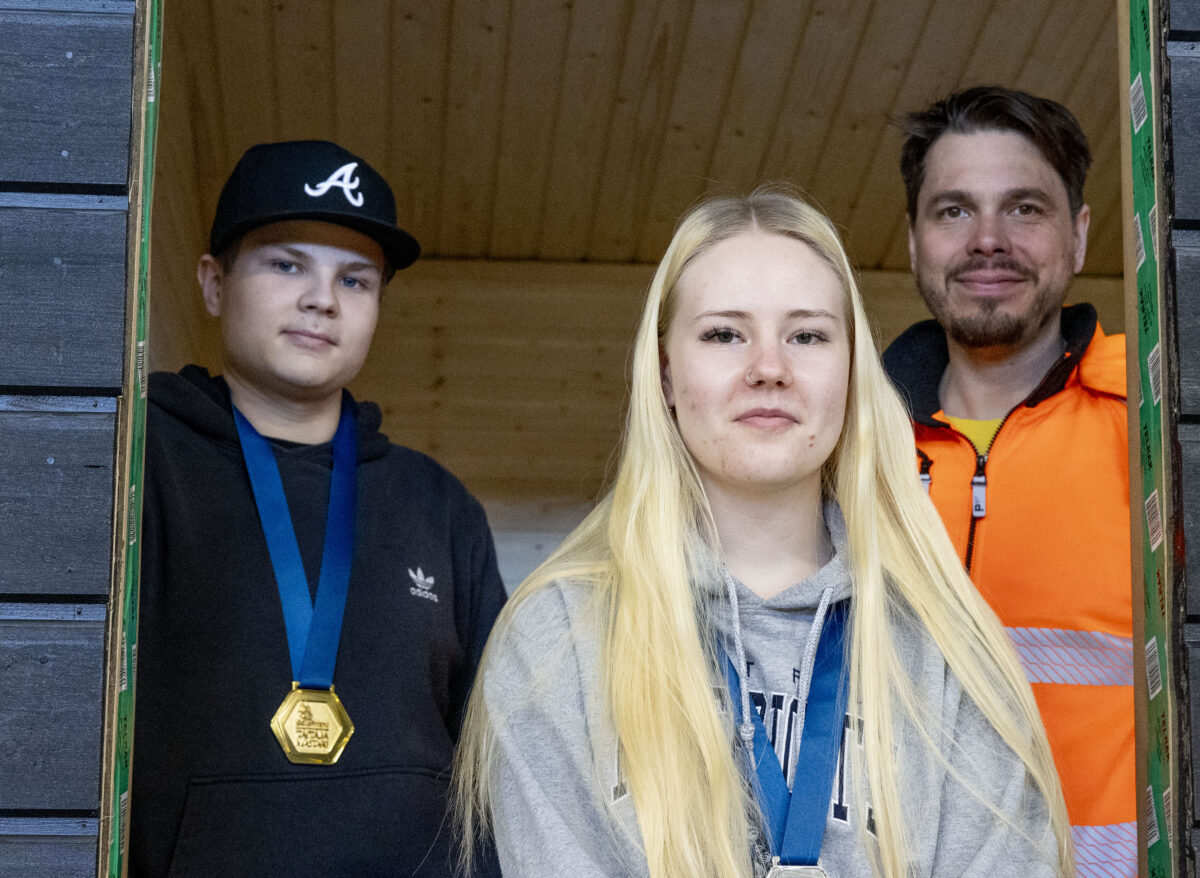 Kaksi opiskelijaa ja opettaja katsovat kameraan rakennuksen ikkunakehikosta.