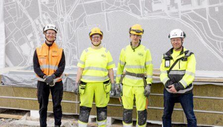 Rakennustyömaan edessä neljä ihmistä rivissä katsomassa kameraan.