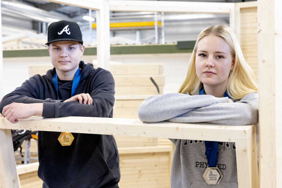 Kaksi ihmistä nojaa puusta tehtyyn kehikkoon ja katsoo kameraan.