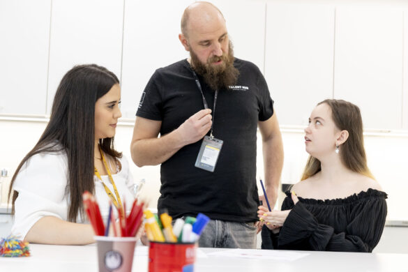Ohjaaja TImo neuvoo kahta opiskelijaa luokkatilassa. Opettaja seisoo keskellä ja keskustelee opiskelijoiden kanssa, jotka istuvat pöydän ääressä. Pöydällä on värikyniä ja toimistotarvikkeita.