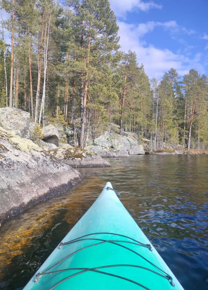 Kajakin keula vedessä kallioisen ja metsäisen rannan edustalla. Taustalla näkyy mänty- ja koivupuita kirkkaan sinistä taivasta vasten.
