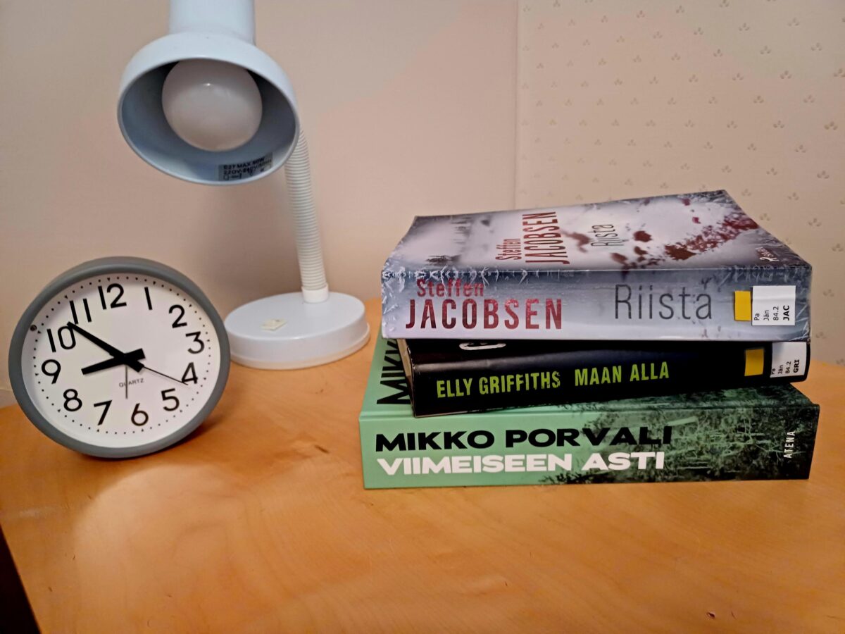 Pöydällä valkoinen pöytälamppu, pyöreä herätyskello, joka näyttää ajan noin 9.50, ja kolme päällekkäin pinottua kirjaa: Steffen Jacobsenin 'Riista', Elly Griffithsin 'Maan alla' ja Mikko Porvalin 'Viimeiseen asti'.