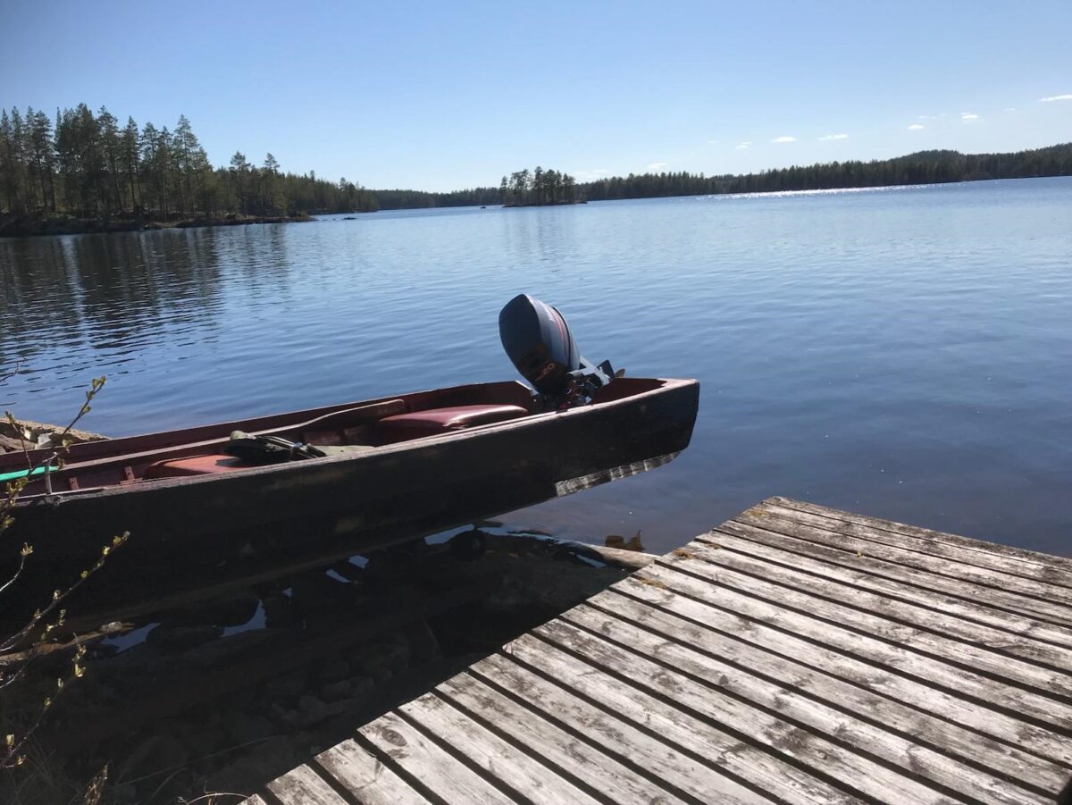 Moottorivene rannassa puisella laiturilla kirkasvetisen järven äärellä. Taustalla näkyy metsäinen ranta ja sininen taivas.